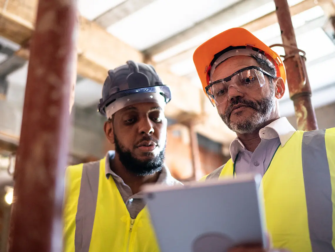 Two blue collar workers wearing hardhats and looking at a tablet together.