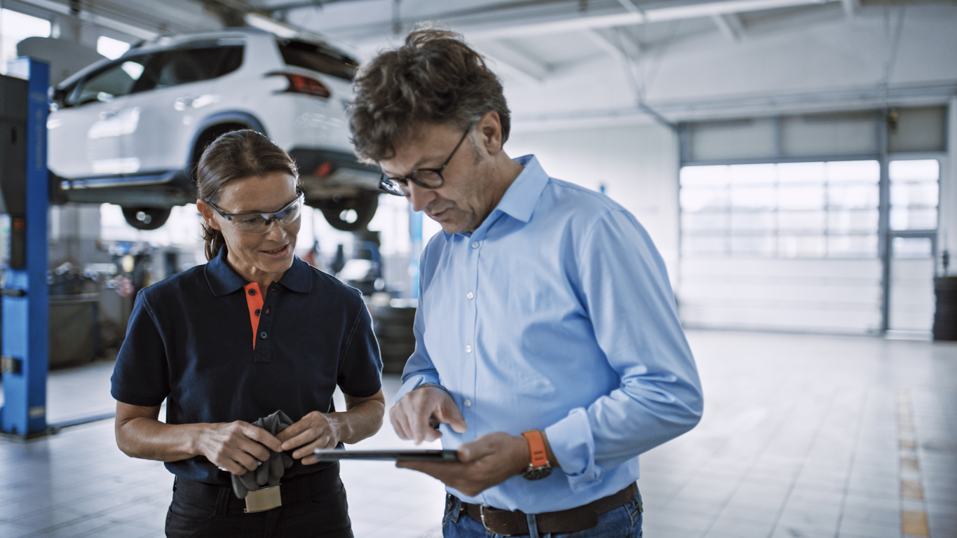 Auto Repair Shop Manager Using Digital Tablet And Having Discussion With A Skilled Woman Mechanic At Workshop.